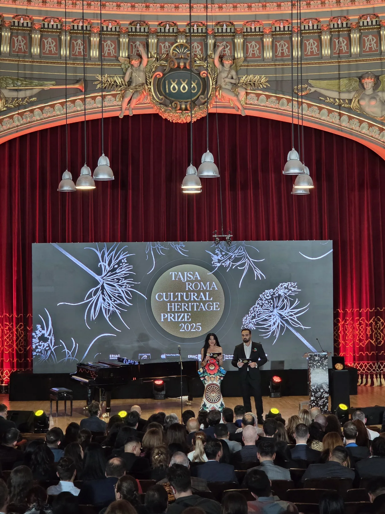 Speaker on stage in front of event graphics at the European Roma Institute programme in the Romanian Athenaeum.